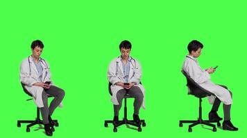 General practitioner texting messages on mobile phone, killing time while she is waiting for patients to arrive at checkup. Woman physician sitting on a chair against greenscreen backdrop. Camera A. video