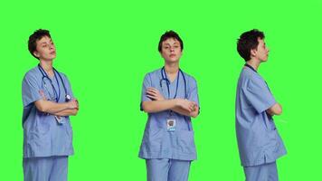 retrato do sorridente médico assistente poses com braços cruzado, mostrando confiança vestido dentro azul hospital esfrega. bem sucedido enfermeira em pé contra tela verde pano de fundo, saúde especialista. Câmera b. video