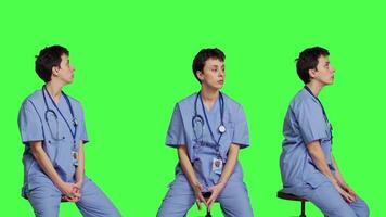 Medical assistant waiting for someone while she sits on chair, feeling impatient and waits for patients to attend checkup appointments. Nurse in scrubs against greenscreen backdrop. Camera B. video