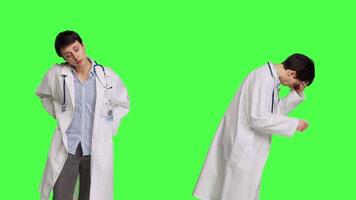 Medic in white coat suffering from migraine and lower back pain, feeling overworked and stressed at hospital. Unwell doctor with headache and spine discomfort against greenscreen backdrop. Camera B. video