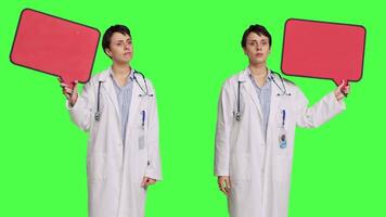 Woman doctor holding a red empty speech bubble sign in studio, showing an isolated blank cardboard icon against greenscreen backdrop. Physician creating announcement with banner. Camera B. video