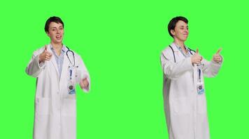 Cheerful medic doing thumbs up symbol against greenscreen backdrop, expresses positivity with like sign. General practitioner with coat giving approval and being satisfied with success. Camera B. video