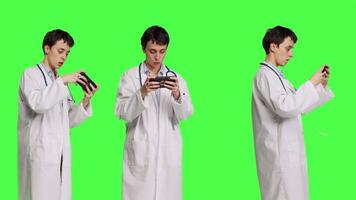 General practitioner playing video games on mobile phone app, having fun with online gaming competition against greenscreen backdrop. Cheerful doctor relaxing with internet game. Camera B.