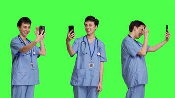 verpleegster beantwoorden video-oproep vergadering verbinding Aan werk pauze, gebruik makend van smartphone naar aansluiten met vrienden Aan online videoconferentie chatten. specialist in scrubs staat tegen groene scherm. camera b. video