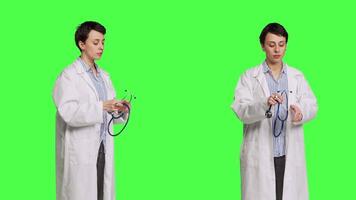 General practitioner with a white coat using stethoscope in studio, preparing to examine patients at checkup appointments. Female doctor in uniform standing against greenscreen backdrop. Camera B. video