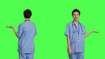 Medical assistant pointing at something aside and doing web commercial, creating advertisement against greenscreen backdrop. Young nurse with scrubs showing a thing to left or right sides. Camera B. video