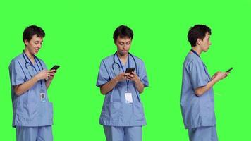 Young nurse texting on smartphone against greenscreen backdrop, using mobile phone to check social media messages on work break. Medical assistant browsing online web pages. Camera B. video