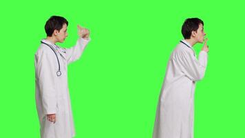 General practitioner showing mute hush sign and calling people to come closer, standing in white coat against greenscreen backdrop. Physician asking to be quiet, inviting someone over. Camera B. video