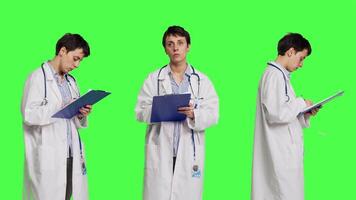 Physician taking notes after patient examination against greenscreen backdrop, writing down medicaments to cure illness. Doctor in white coat using clipboard papers for checkup. Camera B. video