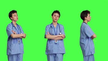 Portrait of confident medical assistant posing with arms crossed in studio, standing against greenscreen backdrop. Successful nurse with healthcare expertise wearing blue scrubs uniform. Camera B. video