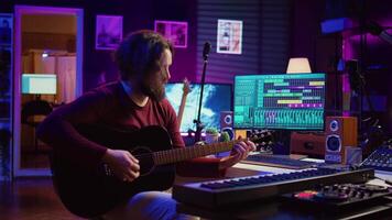 Portrait of male songwriter playing acoustic guitar and recording musical sounds to use for mix and master in home studio. Composing tunes with digital audio workstation software. Camera B. video
