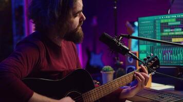 Songwriter producing tunes on acoustic guitar in his personal studio, using a microphone to sing the chords. Audio engineer works with soundboard and equalizer to compose new music. Camera A. video