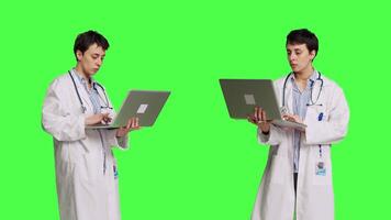 Confident physician searching for new medicaments to treat disease, using laptop to browse on online web pages against greenscreen backdrop. Medic in white coat checks internet sites. Camera B. video