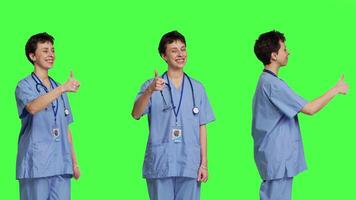 Cheerful nurse giving thumbs up sign against greenscreen backdrop, showing positive gesture with a like. Medical assistant shows her approval, recommending something and being satisfied. Camera B. video