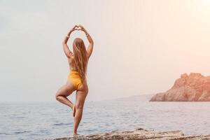 mujer mar yoga. espalda ver de gratis calma felicidad satisfecho mujer con largo pelo en pie en parte superior rock con yoga posición en contra de cielo por el mar. sano estilo de vida al aire libre en naturaleza, aptitud concepto. foto