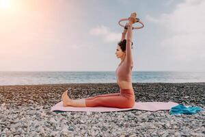 mujer mar pilates deportivo contento medio Envejecido mujer practicando aptitud en playa cerca mar, sonriente activo hembra formación con anillo en yoga estera afuera, disfrutando sano estilo de vida, armonía y meditación foto