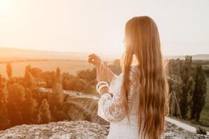 Happy woman in white boho dress on sunset in mountains. Romantic woman with long hair standing with her back on the sunset in nature in summer with open hands. Silhouette. Nature. Sunset. photo