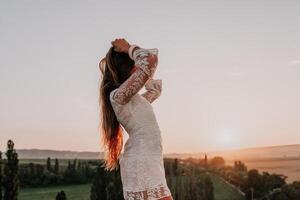 Happy woman in white boho dress on sunset in mountains. Romantic woman with long hair standing with her back on the sunset in nature in summer with open hands. Silhouette. Nature. Sunset. photo