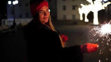 contento sorridente donna nel un' rosso cappello sta su strada, Tenere sparklers nel sua mani. Bengala luce, festivo feyerferk. inverno divertimento Natale o nuovo anno celebrazione. lento movimento, selettivo messa a fuoco video