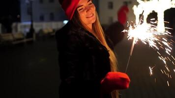 Lycklig leende kvinna i en röd hatt står på gata, innehav sparklers i henne händer. bengal ljus, festlig feyerferk. vinter- roligt jul eller ny år firande. långsam rörelse, selektiv fokus video