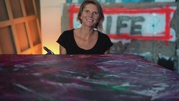 a woman smiling while sitting at a table with a painting on it video