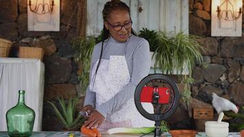 africano Senior mulher preparando Comida receita enquanto transmissão conectados com Móvel Smartphone cam para rede cozinha classe mestre canal video