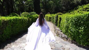 gelukkig vrouw rennen in de park tussen weelderig groen struiken, slow-motion. meisje in wit vliegend jurk met lang blond haar- in de tuin. slank dames- buitenshuis lente vreugde video