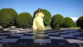 contento donna camminare e gira su scacchiera nel il parco tra lussureggiante verde cespugli, rallentatore. ragazza nel giallo volante vestito con lungo capelli nel il giardino. sottile della signora all'aperto primavera gioia video