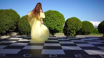gelukkig vrouw wandelen en spins Aan schaakbord in de park tussen weelderig groen struiken. meisje in geel vliegend jurk met lang haar- in de tuin. slank dames- buitenshuis lente vreugde video