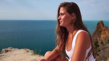 mulher viagem retrato. feliz mulher com grandes cabelo olhando às Câmera e sorridente, partilha dela viagem histórias com amigos. fechar acima retrato mulher posando em uma vulcânico Rocha Alto acima mar. lento movimento video