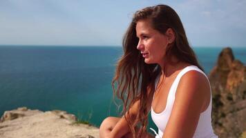 Woman travel portrait. Happy woman with long hair looking at camera and smiling, sharing her travel stories with friends. Close up portrait woman posing on a volcanic rock high above sea. Slow motion video