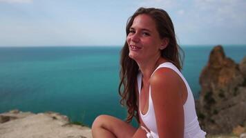 Woman travel portrait. Happy woman with long hair looking at camera and smiling, sharing her travel stories with friends. Close up portrait woman posing on a volcanic rock high above sea. Slow motion video