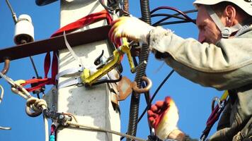 Skilled electrician in helmet fixes wires standing on ladder near high pole against blue sky on summer day backside view. Electrical service and mounting on the pole. Slow motion video