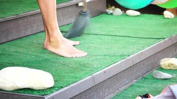 unrecognizable man sweeping the green carpet of artificial lawn on the beach from pebbles with broom. video