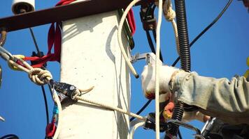 Skilled electrician in helmet fixes wires standing on ladder near high pole against blue sky on summer day backside view. Electrical service and mounting on the pole. Slow motion video