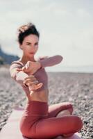 Middle aged well looking woman with black hair, fitness instructor in leggings and tops doing stretching and pilates on yoga mat near the sea. Female fitness yoga routine concept. Healthy lifestyle photo