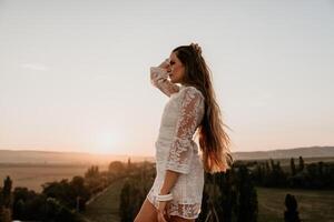 Happy woman in white boho dress on sunset in mountains. Romantic woman with long hair standing with her back on the sunset in nature in summer with open hands. Silhouette. Nature. Sunset. photo