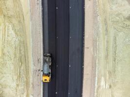 yellow truck pouring bitumen over a new road. Aerial drone shot. Road construction in progress on slope nature canyon. Infrastructure development and logistics. photo