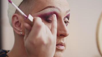 teatro masculino ator aplica-se Rosa sombras em a pálpebras e misturas eles enquanto Maquiagem com uma mão espelho dentro a vestir quarto fechar acima durante preparação para a desempenho video