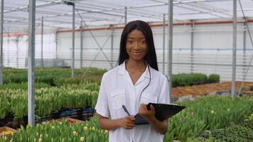 africano americano jovem menina florista ou nerd dentro uma branco camisa carrinhos em uma fundo do linhas do tulipa mudas com uma caneta e uma tábua dentro dele mãos video