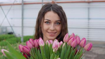 portret van een blij jong vrouw bloemist in een rood en zwart overhemd met een boeket van roze tulpen in haar handen. arbeider in de kas video