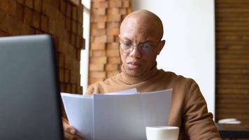 un africano americano hombre con un grave expresión trabajos con un ordenador portátil y documentos a hogar. pandemia. encierro video