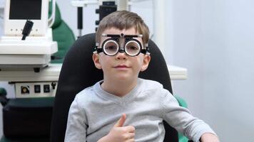 pequeño chico en un juicio marco para un selección de lentes es sentado en un óptica Tienda video