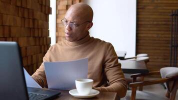 uma homem com uma sério expressão trabalho com uma computador portátil e documentos dentro uma cafeteria. caramelo estética. pandemia video