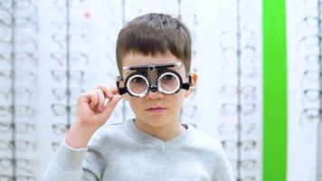 wenig Junge steht auf ein Hintergrund von ein Schaufenster mit Frames zum Brille mit das Gerät zum Auswahl von Kontakt Linsen video