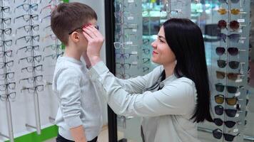 Young caucasian woman with baby, mother and son choosing glasses in optics store video