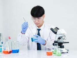 retrato asiático hombre estudiante científico médico vestido uno persona sti en laboratorio mirando mano participación prueba tubo químico. causado por mezcla reactivos en científico investigación laboratorios y microscopio en mesa foto