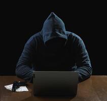 Portrait hacker spy man one person in black hoodie sitting on table looking computer laptop used login password attack security to data digital internet network system night dark background copy space photo