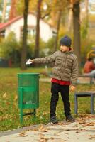 lleno longitud retrato de un seis año antiguo chico lanzamiento papel a el basura compartimiento basura lata en el otoño parque foto