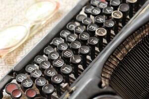 Black vintage metal type writer next to a pile of old note books, pair of eyeglasses and a red pencil on a linen tablecloth photo